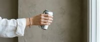 Woman holding silver aluminum can beverage drink in hand wearing white shirt sleeve with bracelet against neutral gray wall background for lifestyle marketing.