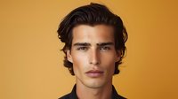 Young Hispanic male model with dark wavy hair and defined facial features poses confidently against warm orange background in professional studio portrait.
