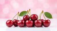 Fresh ripe red cherries with green leaves arranged in row on white surface against soft pink bokeh background for summer fruit concepts.
