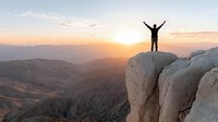 Silhouette of triumphant hiker with raised arms standing on rocky mountain peak at golden sunrise overlooking vast valley landscape for motivation concepts.