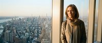 Young professional woman in beige coat standing by floor-to-ceiling windows overlooking urban cityscape at sunset, contemplating business opportunities and career success.