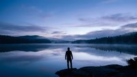 Silhouette of man standing on rocky shore at peaceful mountain lake during twilight with perfect water reflections and misty forest landscape.