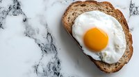 Fresh sunny side up egg on rustic sourdough bread slice against marble background, minimalist breakfast composition with vibrant yolk and white edges.