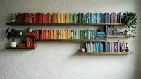 Colorful books arranged by rainbow spectrum on floating wooden shelves against white brick wall with plants creating organized home library display.
