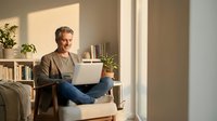 Mature Caucasian man working on laptop computer at home in bright living room with natural sunlight streaming through window creating warm atmosphere.