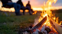 Glowing campfire with burning logs and flames at sunset, silhouettes of people relaxing in background on grassy field during outdoor camping adventure.