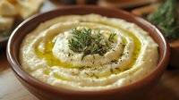 Creamy hummus topped with olive oil, fresh rosemary herbs and black pepper in traditional ceramic bowl, close up view of middle eastern appetizer dip.