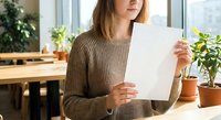 Young Caucasian woman in brown sweater reading document at home office desk with plants. Remote work, paperwork review, business correspondence concept.