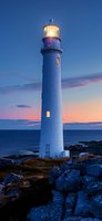 White lighthouse illuminated at dusk against dramatic purple and orange sunset sky, standing tall on rocky coastal cliff overlooking calm ocean waters.