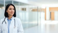 Professional Hispanic female doctor in white coat with stethoscope standing confidently in modern hospital corridor for healthcare marketing materials.