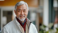 Senior Asian man with gray beard wearing white winter coat and scarf smiling warmly at camera, expressing joy and confidence against blurred urban background.