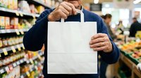Man holding blank white paper shopping bag in grocery store with stocked shelves and other shoppers in background, suitable for retail marketing and consumer advertising.