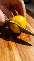 Hand slicing fresh yellow lemon in half on wooden cutting board with sharp knife, food preparation and cooking ingredient.