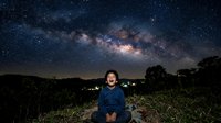 Young adult male with light skin tone sitting outdoors at night, gazing upward at starry sky and Milky Way galaxy. Suitable for astronomy, inspiration, and wonder concepts.