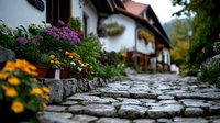 Stone pathway through traditional European village with colorful flower beds of purple and yellow blooms along white cottage houses, creating charming rustic atmosphere.