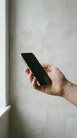 Male hand holding a modern smartphone with blank black screen against a light textured wall, mockup for app or mobile design.