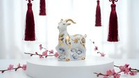 Young woman with light skin tone and blonde hair, wearing elegant white and gold attire, posing on white cylindrical platform surrounded by decorative burgundy tassels and pink cherry blossoms for luxury fashion and lifestyle content.