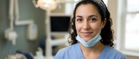 Female healthcare professional with dark wavy hair and medium complexion wearing blue medical scrubs and face mask in clinical setting. Portrait for medical, nursing, and healthcare industry use.