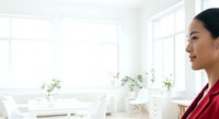 Asian woman in red blazer working at bright, modern office desk with multiple windows and potted plants, professional business environment.