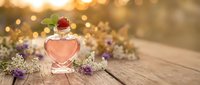 Heart-shaped glass bottle with pink perfume surrounded by lavender flowers on wooden surface with warm golden bokeh lights creating romantic atmosphere.