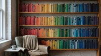 Cozy reading nook with rainbow organized bookshelf displaying colorful books arranged by hue, comfortable armchair with throw blanket near bright window.