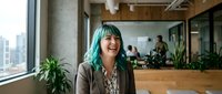 Young Asian woman with turquoise hair laughing in modern office workspace with plants and city view through windows.