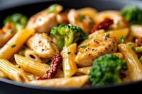Penne pasta with grilled chicken pieces, fresh broccoli florets and sun-dried tomatoes in creamy sauce, garnished with black pepper and herbs, served in dark plate.