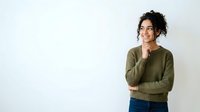 Young woman with curly dark hair and olive skin tone wearing olive green sweater and blue jeans, standing against white background with thoughtful expression, hand near chin, suitable for lifestyle and portrait use.