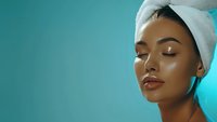 Young Asian woman with glowing skin wearing white towel on head against turquoise background, eyes closed, demonstrating spa and skincare concept.