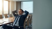 Relaxed businessman leaning back in office chair with hands behind head, smiling confidently in modern conference room during break from work.
