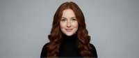 Professional redhead woman with wavy hair wearing black turtleneck sweater smiling confidently against neutral gray background for business portraits.