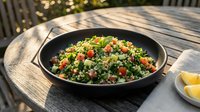 Tabbouleh salad with fresh parsley, tomatoes, and bulgur wheat served in black bowl on outdoor wooden table with lemon wedges.