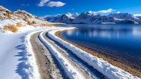 Winding snow-covered mountain road along tranquil blue lake with majestic peaks and white clouds reflecting in crystal clear water on sunny winter day.