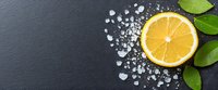 Fresh cut lemon half with green leaves and scattered sea salt crystals on dark slate background, food photography composition with copy space.