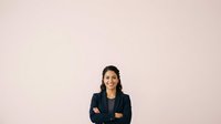 Young woman with dark hair and South Asian ethnicity wearing professional navy blazer and glasses, arms crossed in confident pose against neutral pink background, suitable for corporate, business, and professional website use.