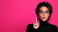 Young Hispanic woman in black turtleneck with glasses making surprised expression and raised finger against bright pink background, messy bun hairstyle. For advertising or blog content.