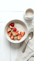 Overhead view of yogurt bowl with fresh strawberries and quinoa seeds on white wooden surface, styled with milk pitcher and spoon for healthy breakfast or wellness content.