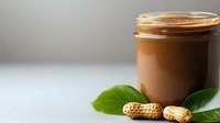 Creamy peanut butter in glass jar with fresh green leaves and raw peanuts on white surface against neutral background, healthy food concept.