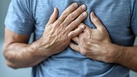 Man in grey t-shirt clutching chest with both hands, showing signs of chest pain, heart attack symptoms, or breathing difficulties. Medical emergency concept.
