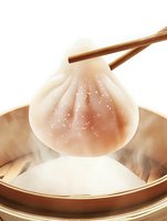 Steaming hot Asian dumpling held by wooden chopsticks over bamboo steamer with rising vapor, showcasing traditional dim sum cuisine in warm lighting.