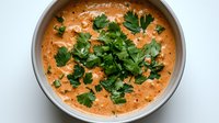 Creamy orange-hued soup garnished with fresh parsley leaves in gray ceramic bowl, overhead view showing rich texture and vibrant herbs against smooth background.