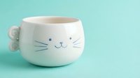 Adorable white ceramic cat face mug with whiskers and smile on turquoise background for kitchen decor and coffee lovers.