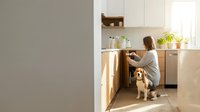 Young woman with brown hair in casual sweater cooking in modern kitchen with golden retriever dog sitting nearby on wooden floor during sunny morning.