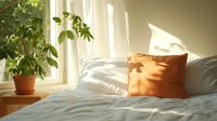 Cozy bedroom interior with morning sunlight casting shadows through sheer curtains, orange accent pillow on white bedding, and green houseplant by window.