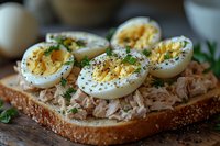 Open-faced sandwich with tuna salad and sliced hard-boiled eggs topped with fresh herbs and black pepper on artisanal bread, macro food photography.