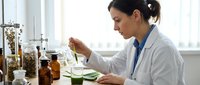 Woman scientist in lab coat conducting herbal extraction experiment with dried plants and glass containers for botanical research, natural medicine development, or pharmaceutical testing.