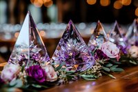 Glass pyramid centerpieces with purple roses and eucalyptus on wooden table, illuminated by warm bokeh lights creating romantic wedding reception atmosphere.