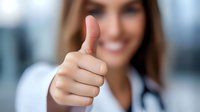 Young female medical professional showing thumbs up gesture with confident smile, wearing white coat and stethoscope in hospital setting. Focus on hand.