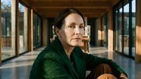 Mature woman with dark hair and fair complexion, wearing a dark green cardigan, sitting thoughtfully in a modern corridor with large windows and wooden ceiling beams, natural daylight streaming through glass panels.