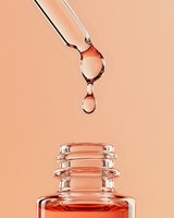 Glass dropper dispensing amber serum drops into open bottle against warm peach background for skincare and cosmetic product applications.